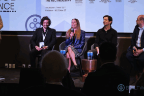 A businesswoman smiles as she speaks into a microphone addressing an audience. Other panelists sit on either side of her.
